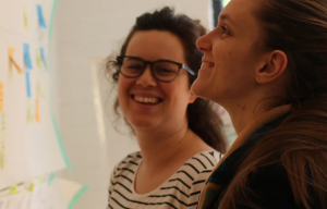 Photo de l'équipe au travail : deux personnes souriantes collaborent face à un mur sur lequel sont affichés des notes
