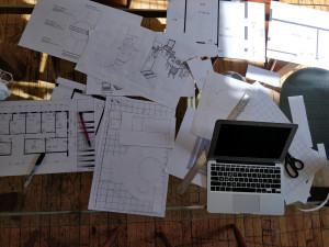photo d'une table de travail encombrée de crocquis, de plans, de feuilles de notes, d'un ordinateur, de crayons et d'une règle graduée