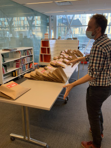 Photographie d'une personne debout devant une table, où est posé plusieurs sacs, dans une bibliothèque.