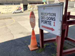 Au premier plan, un panneau avec le logo de Estrie Aide sur lequel il est écrit “Réception des dons, porte 1”. À sa gauche, un cône orange dans lequel est planté un panneau rouge et blanc “Arrêt”. Au second plan, une voiture blanche est stationnée de l’autre côté de la rue, devant une murale qui inclut le message “On se ressemble plus qu’on ne le pense”.