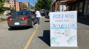 Au premier plan de la photo, un tableau blanc sur lequel est écrit en grandes lettres bleues “Aidez-nous à mieux vous accueillir” invite à participer à une enquête. Au second plan, une femme en chemise blanche et en jean se penche vers une voiture grise pour demander à son conducteur si il souhaite participer.