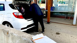 Au premier plan de la photo, du papier et un crayon placés sur une tablette pour la prise de note sont posés sur un muret en béton. Au second plan, un couple de donateurs déchargent des dons de leur voiture blanche.