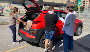 Au premier plan de la photo, deux femmes échangent tandis qu’au deuxième plan, une troisième femme décharge des articles d’une voiture rouge.