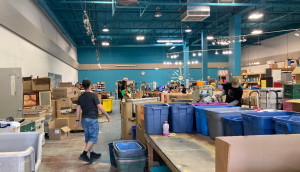 Photo d’une grande salle munie de tables et de boîtes en plastique bleues dans lesquels des articles donnés sont triés. Au premier plan, un homme en t-shirt noir et portant une casquette se dirige vers l’arriére de la salle.